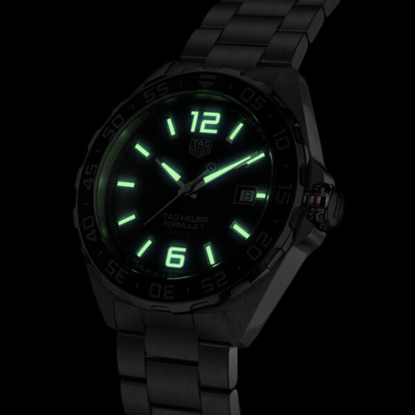 Eine schwarze Armbanduhr mit leuchtend grünem Zifferblatt ist vor einem dunklen Hintergrund zu sehen. Die Uhr hat leuchtende Zeiger und Markierungen und ein Metallarmband. Sie zeigt das Datum an und auf dem Zifferblatt steht „TAG Heuer Formula 1“.