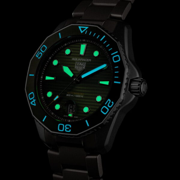 Eine schwarze Tag Heuer Aquaracer-Uhr mit leuchtendem Zifferblatt und leuchtenden Zeigern. Die grün und blau leuchtenden Markierungen und Zahlen heben sich vom dunklen Zifferblatt und Hintergrund ab und zeigen Zeitmarkierungen und Logo.