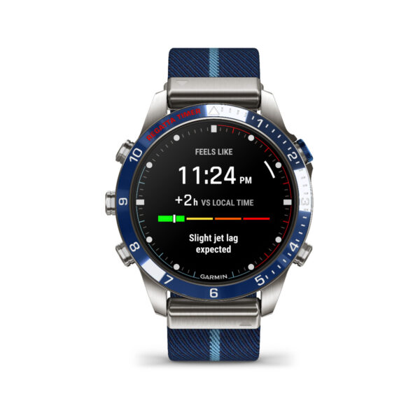 Die Smartwatch Garmin Marq Captain 2.Gen. mit blauer Lünette und gestreiftem Armband zeigt Uhrzeit und Datum mit „Fühlt sich an wie 23:24 Uhr“ und „+2 Std. gegenüber Ortszeit“ an, zusammen mit einer leichten Jetlag-Warnung.