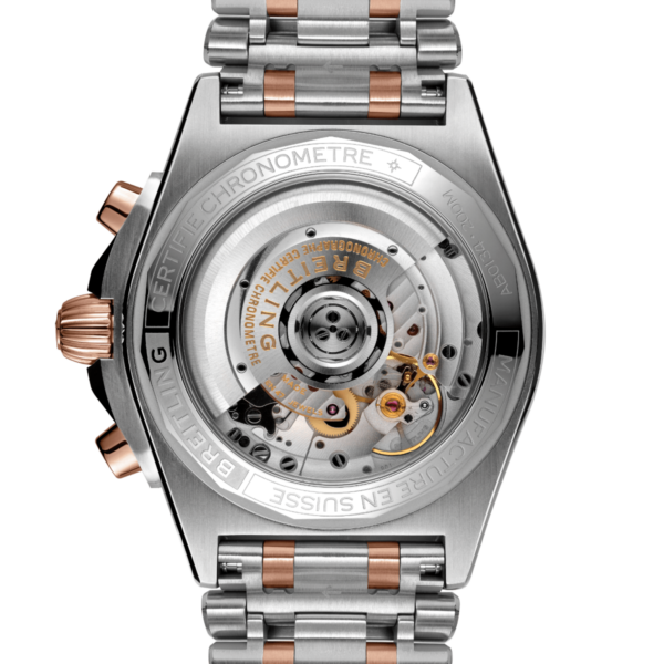 Eine Nahaufnahme zeigt das komplexe Uhrwerk im transparenten Gehäuseboden des Breitling Chronomat B01 Chronograph 42. Die Uhr verfügt über ein Metallarmband in Roségold- und Silbertönen, auf dessen Gehäuseboden deutlich sichtbar die Aufschrift „Chronometre“ und „Breitling“ zu sehen ist.