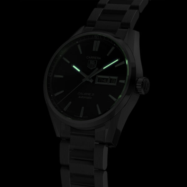 Die TAG Heuer Carrera ist eine dunkel getönte Armbanduhr mit leuchtend grünen Markierungen und Zeigern, einem Metallarmband und einer Tages-/Datumsanzeige. Der Markenname ist auf dem Zifferblatt neben „Calibre 5 Automatic“ zu sehen.