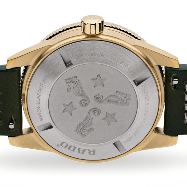 Die Rückseite der Rado Captain Cook Automatic Bronze-Armbanduhr hat ein Bronzegehäuse und ein grünes Lederarmband. Auf der Metallrückseite des Gehäuses sind drei Seepferdchen und fünf Sterne mit umlaufendem Text eingraviert. Auf dem Armband ist eine Seriennummer angebracht.