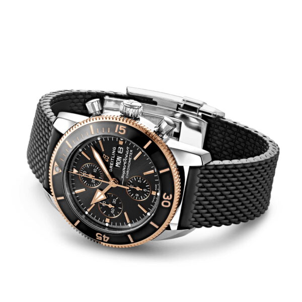 Die Breitling Superocean Héritage II 44 ist eine Luxusarmbanduhr mit schwarzem Zifferblatt und drei Hilfszifferblättern, goldenen und silbernen Akzenten sowie einem schwarzen strukturierten Armband. Die Lünette verfügt über Minutenanzeigen, und zwei Drücker rechts neben der Krone sorgen für ein elegantes Erscheinungsbild.