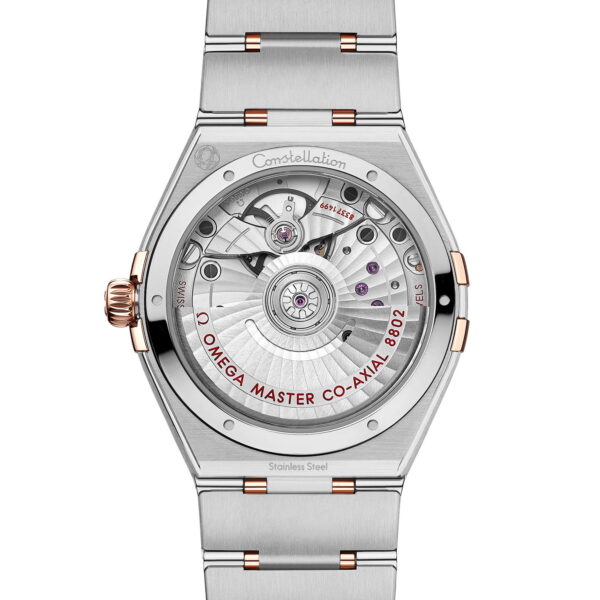 Die Rückseite einer OMEGA Constellation Small Seconds 34 mm präsentiert eine silberne Armbanduhr mit Edelstahlarmband und gibt den Blick auf das mechanische Uhrwerk frei. Sie ist mit Roségold-Akzenten versehen und trägt die Gravur „OMEGA MASTER CO-AXIAL 8902“ um den Mechanismus.