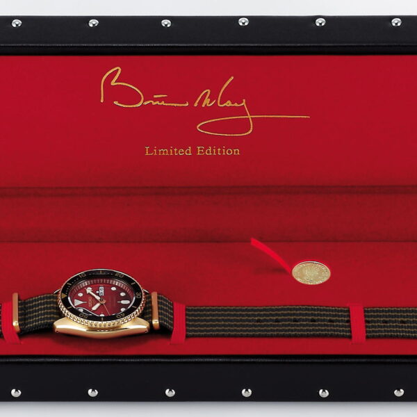 Die Seiko 5 Sports Brian May Limited Edition, eine gold-schwarze Uhr mit rotem Zifferblatt, ruht in einem offenen schwarz-roten Gehäuse mit silbernen Nieten. Das Gehäuseinnere ist rot und trägt den goldenen Schriftzug „Limited Edition“. Neben der Uhr befindet sich eine kleine Goldmünze.
