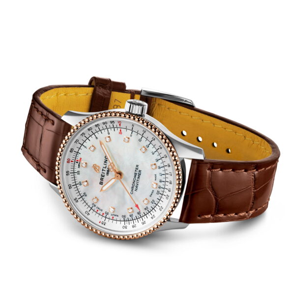 Die Breitling Navitimer Automatic 35 besticht durch ein braunes Lederarmband, eine polierte Lünette aus Silber und Gold sowie ein weißes Zifferblatt mit roten und schwarzen Markierungen, Ziffern und Chronographendetails unter transparentem Glas. Das Armband ist mit einer Faltschließe ausgestattet.
