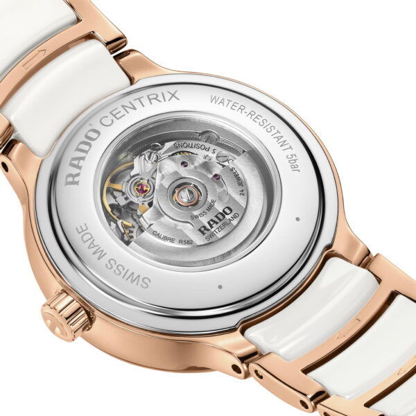 Eine Nahaufnahme zeigt die Rückseite der Rado Centrix Automatic Diamonds mit ihrem freiliegenden mechanischen Uhrwerk. Sie verfügt über ein Armband aus Roségold und Weiß mit den Aufschriften „Water-Resistant 5 bar“, „Swiss Made“ und „Rado“.