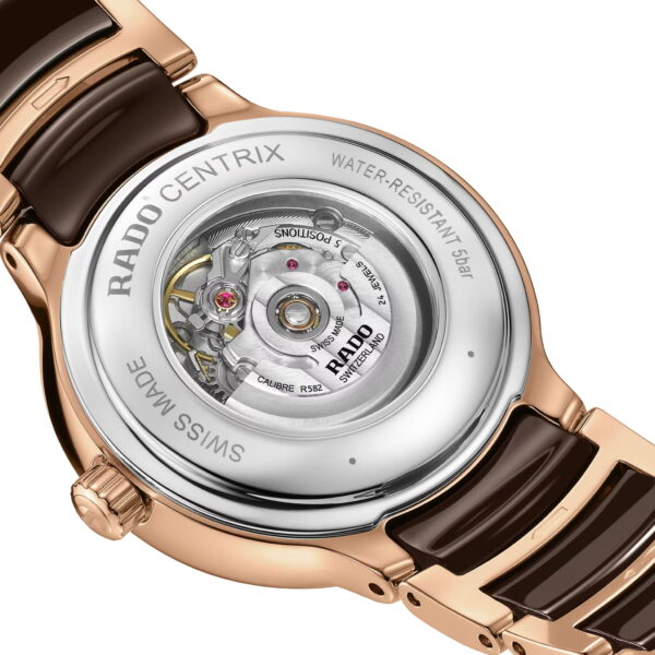 Nahaufnahme der Rückseite einer Rado Centrix Automatic Diamonds. Das transparente Gehäuse gibt den Blick auf die komplexe Mechanik frei. Das Armband besteht aus goldfarbener und schwarzer Keramik. Der gravierte Rand weist auf die Swiss Made-Herkunft und Wasserdichtigkeit hin.