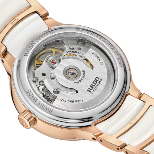 Die luxuriöse Rado Centrix Automatic Diamonds präsentiert ihr komplexes mechanisches Uhrwerk durch einen transparenten Gehäuseboden. Das Gehäuse besteht aus Edelstahl und Roségold und wird von einem weißen Keramikarmband getragen. Auf der Rückseite sind der Markenname und die technischen Daten eingraviert.