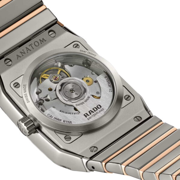 Die Nahaufnahme der Rado Anatom Automatic offenbart ihr mechanisches Uhrwerk. Der sichtbare Mechanismus ist mit technischen Daten beschriftet und durch eine transparente Abdeckung geschützt. Das Metallarmband ist in Silber und Roségold gehalten.