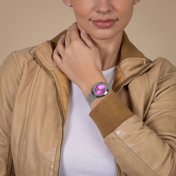 Sie tragen eine hellbraune Lederjacke und ein weißes Oberteil und zeigen die Rado DiaStar Original Automatic-Uhr mit leuchtend rosa Zifferblatt, die sanft ihren Hals vor einem beigen Hintergrund berührt.