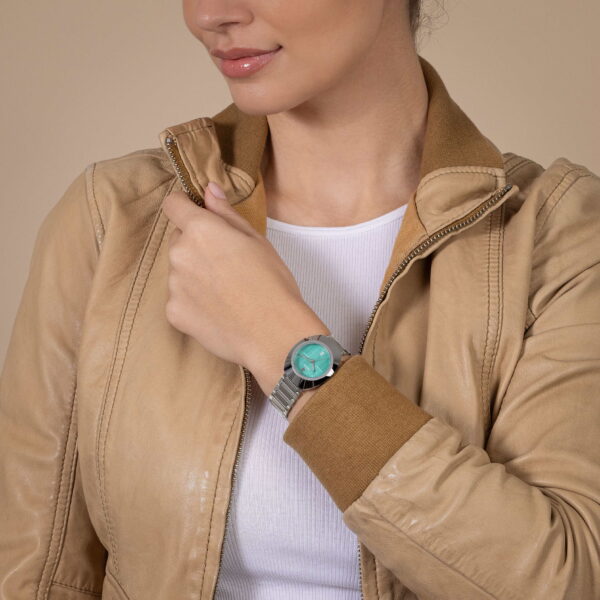 Eine Person trägt eine hellbraune Lederjacke über einem weißen Hemd und präsentiert die Rado DiaStar Original Automatic mit türkisfarbenem Zifferblatt. Sie richtet mit einer Hand ihren Kragen und gibt so einen Teil ihres Gesichts vor einem neutralen beigen Hintergrund frei.