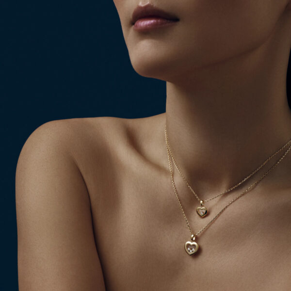 Eine Person mit nackten Schultern trägt zwei Chopard Happy Diamonds Collier Goldketten mit herzförmigen Anhängern. Der tiefblaue Hintergrund hebt den Schmuck auf ihrer Haut hervor.