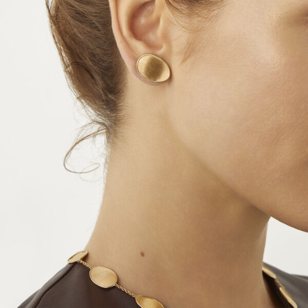 Eine Person mit heller Haut trägt goldene ovale Ohrstecker „Lunaria“ von Marco Bicego und eine passende goldene Halskette mit flachen ovalen Gliedern. Ihr Haar ist zurückgebunden, und sie trägt ein braunes Oberteil. Nur die Seite ihres Gesichts ist sichtbar.