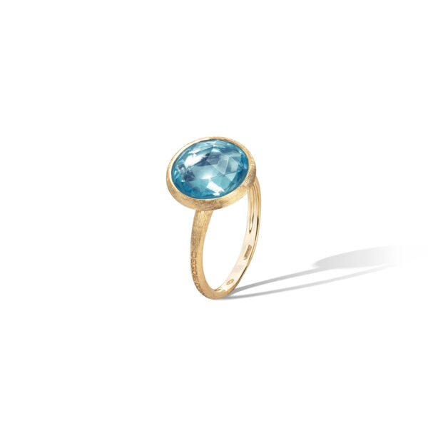 Ein goldener Ring mit einem großen, runden blauen Topas-Edelstein in einer einfachen Zargenfassung wirft einen weichen Schatten auf einen weißen Hintergrund.