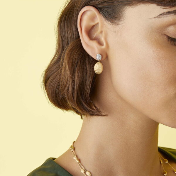 Nahaufnahme eines Frauenohrs mit den Marco Bicego Siviglia Ohrhängern, goldenen Ohrringen mit runden, strukturierten Anhängern und einem kleinen Diamanten. Sie trägt außerdem eine passende goldene Halskette, hat kurze Haare und der Hintergrund ist in zartem Gelb gehalten.