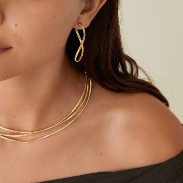 Eine Frau trägt ein schwarzes schulterfreies Top, goldene Creolen-Ohrringe von Marco Bicego Marrakech und eine dreireihige Goldkette. Der Schmuck sticht vor ihren nackten Schultern und ihrem langen braunen Haar hervor.
