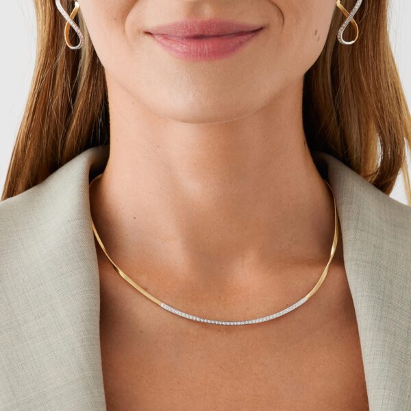 Eine Frau mit langen Haaren und einem hellgrauen Blazer präsentiert das Marco Bicego Collier Marrakech, das eine goldene Halskette mit kleinen Diamanten und passende goldene Ohrringe umfasst. Ihre leicht rosa Lippen und ihr Blick auf den Hals sind vor einem weißen Hintergrund zu sehen.