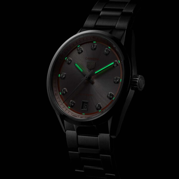 Die TAG Heuer Carrera Date ist eine schwarze Metallarmbanduhr mit dunklem Zifferblatt, leuchtend grünen Stunden- und Minutenzeigern, Punktstundenmarkierungen und einem Datumsfenster bei 6 Uhr, das vor einem schwarzen Hintergrund angezeigt wird.