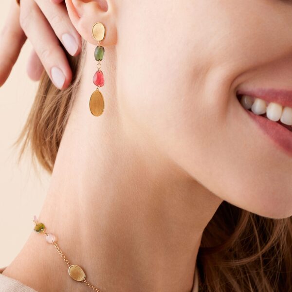 Eine Nahaufnahme einer lächelnden Frau, die Marco Bicego Lunaria Creolen trägt – lange goldene Ohrringe mit ovalen Elementen in Grün, Rosa und Gold – und ein passendes Armband mit rosa und goldenen Akzenten.