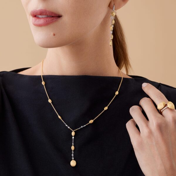Eine Frau in einem schwarzen Oberteil präsentiert Goldschmuck, darunter die Marco Bicego Siviglia Kette mit Goldperlen, einen baumelnden Ohrring und einen Goldring. Ihr Haar ist zurückgebunden, während sie ihre Hand in Richtung ihres Schlüsselbeins hebt.