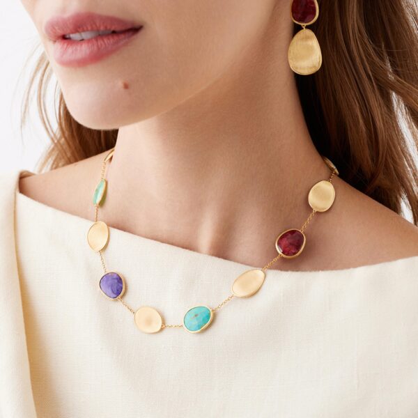 Nahaufnahme einer Frau in einem cremefarbenen Oberteil. Sie trägt die Marco Bicego Lunaria Kette, eine goldene Halskette mit ovalen bunten Edelsteinen, sowie passende goldene Ohrringe. Ihr offenes Haar bedeckt teilweise ein Ohr.