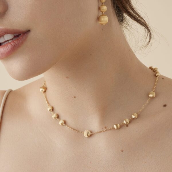Nahaufnahme einer Frau mit heller Haut und geöffneten Lippen. Sie trägt die Marco Bicego Africa Kette, eine zarte Halskette aus Goldperlen, und dazu passende goldene Ohrhänger. Ihr Haar ist zurückgebunden und sie trägt ein dünnes Trägertop.
