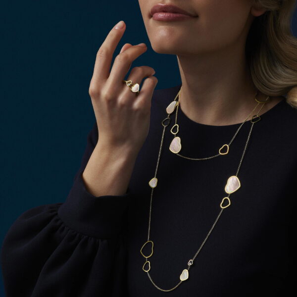 Eine Frau in einem dunklen Langarmshirt trägt die mehrlagigen Goldketten und den Ring von Perlmutt/Brillant, jeweils mit unregelmäßigen blassrosa Steinen besetzt, vor einem dunkelblauen Hintergrund. Ihr Gesicht ist teilweise sichtbar.