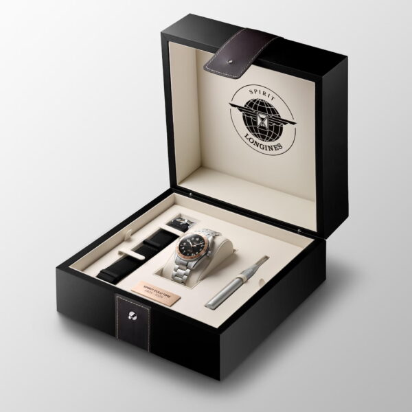 Eine geöffnete schwarze Longines-Box präsentiert die Edelstahluhr Longines Spirit Zulu Time 1925, ein zusätzliches schwarzes Armband, ein Werkzeug zum Armbandwechsel und ein Namensschild. Der Innendeckel trägt das Longines Spirit-Logo.