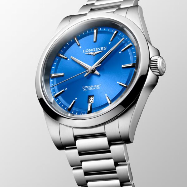 Die Longines Conquest ist eine Armbanduhr aus Edelstahl mit leuchtend blauem Zifferblatt, silbernen Indexen und Zeigern, einer Datumsanzeige bei 6 Uhr und einem Gliederarmband aus Metall, das sich vor einem hellen Hintergrund abhebt.