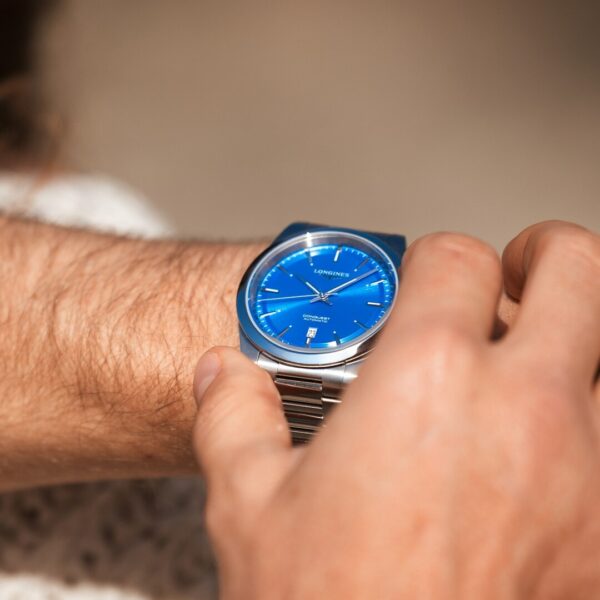 Eine Person stellt eine silberne Longines Conquest-Armbanduhr mit markantem blauem Zifferblatt und silbernen Zeigern an ihrem linken Handgelenk ein, vor einem sanft verschwommenen Hintergrund, der den Zeitmesser hervorhebt.