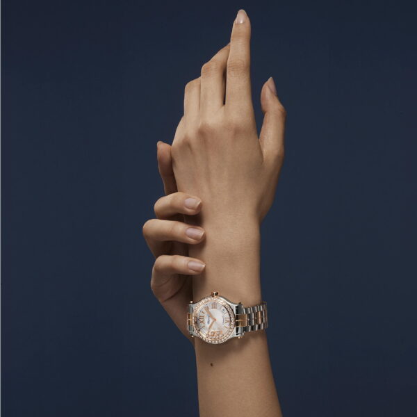 Mit sauber manikürten Fingernägeln präsentiert eine Person die Luxusarmbanduhr Chopard Happy Sport mit Metallband und juwelenbesetzter Lünette vor einem einfarbig dunkelblauen Hintergrund.