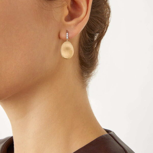 Nahaufnahme eines Frauenohrs, das Marco Bicego Lunaria Ohrhänger trägt, elegante ovale Ohrringe aus Gold mit Edelsteinbesatz. Ihr Haar ist hochgesteckt, sie trägt ein dunkelbraunes Oberteil, und der Hintergrund ist schlicht und hell.