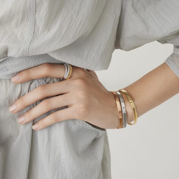 Eine Person, die ein senfgelbes Kleid trägt, hat ihre linke Hand auf die Hüfte gestützt und präsentiert ein silbernes Armband mit eingravierten Details und zwei Ringe an ihrem Ringfinger vor einem neutralen Hintergrund.