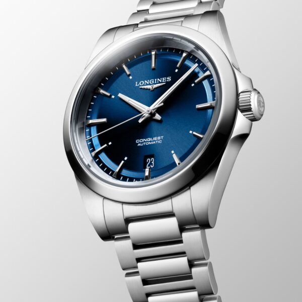 Die Longines Conquest ist eine silberne Automatik-Armbanduhr mit blauem Zifferblatt, Datumsanzeige bei 6 Uhr und einem Edelstahlarmband, das schräg auf einem hellen Farbverlaufs-Hintergrund liegt.