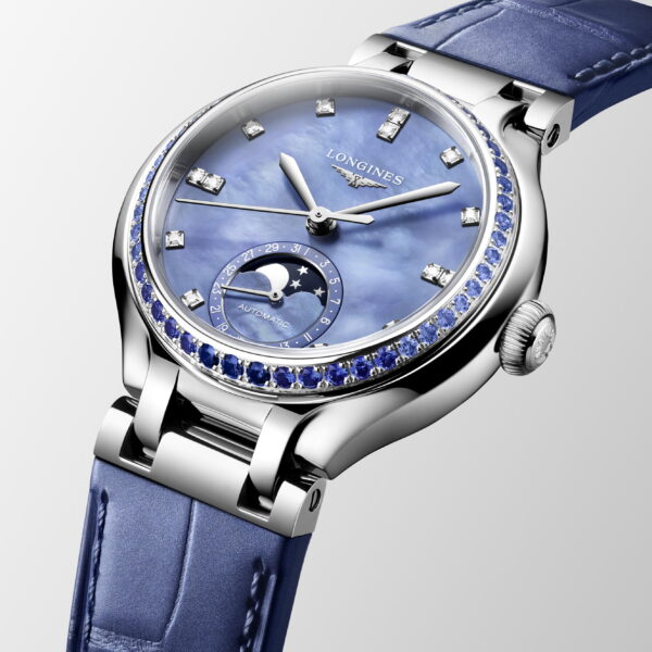 Die Longines Primaluna Moonphase Armbanduhr verfügt über ein blaues Perlmuttzifferblatt, diamantbesetzte Indexe, ein Mondphasen-Zifferblatt, eine Lünette mit blauen Edelsteinen und ein blaues Lederarmband vor einem hellgrauen Hintergrund.