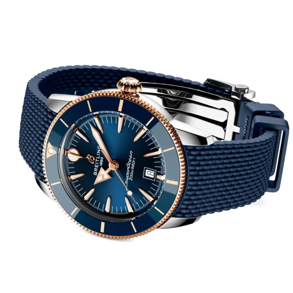 Die Breitling Superocean Heritage B31 Automatic 44 mit blauem Zifferblatt, goldenen Akzenten, Datumsanzeige und blauem Strukturarmband ist schräg auf einem weißen Hintergrund abgebildet.