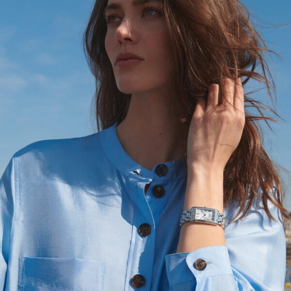 Eine Frau in einer hellblauen geknöpften Bluse steht im Freien vor einem blauen Himmel und schaut weg, während sie mit einer Hand ihr Haar berührt und die silberne Longines DolceVita-Armbanduhr an ihrem Handgelenk zur Schau stellt.