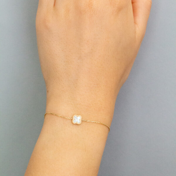 Eine Hand, die ein zartes Goldarmband mit einem kleinen, runden, weißen Edelstein und einen passenden Goldring mit einem ähnlichen Edelstein trägt, dargestellt vor einem schlichten grauen Hintergrund.