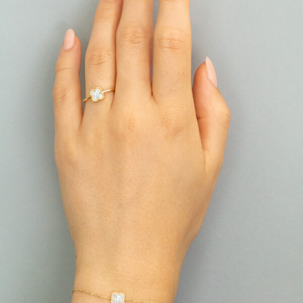 Eine Hand mit manikürten Nägeln trägt einen zarten Goldring mit einem klaren Edelstein und ein dazu passendes Goldarmband mit einem ähnlichen Edelstein, die vor einem schlichten grauen Hintergrund präsentiert werden.