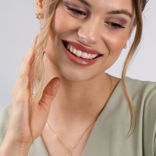 Eine Frau mit hellbraunem Haar in einem hellgrünen Oberteil lächelt, während sie ihr Haar berührt. Sie trägt goldene Ohrringe und das Echtgold Collier mêler, eine goldene Halskette mit einem kleinen Anhänger.