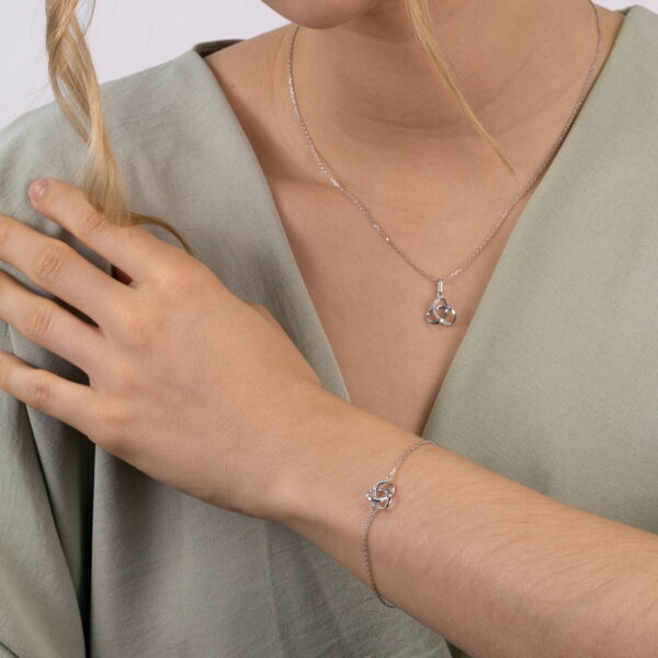 Eine Frau in einem hellgrünen Oberteil präsentiert das Echtgold Armband mêler, ein silbernes Armband mit einem Dreifaltigkeitsknotenmuster, das zu ihrer Halskette passt. Sie berührt ihre Schulter, während eine lockere Locke ihr Gesicht umrahmt.