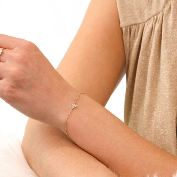Eine Person trägt das Echtgold Armband Effeuiller - ein zartes Goldkettenarmband mit einem kleinen Diamanten in der Mitte - an ihrem Handgelenk. Sie trägt ein ärmelloses beiges Oberteil und einen Ring.