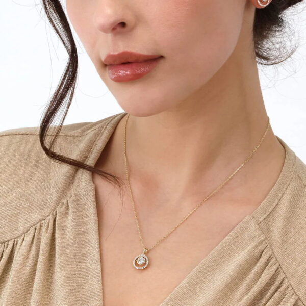 Eine Frau modelliert das Echtgold Collier Cénacle, eine goldene Halskette mit einem runden Anhänger mit einem klaren Edelstein in der Mitte, der von kleineren Steinen umgeben ist. Nur ihr unteres Gesicht, ihr Hals und ihre obere Brust sind vor einem schlichten Hintergrund zu sehen.