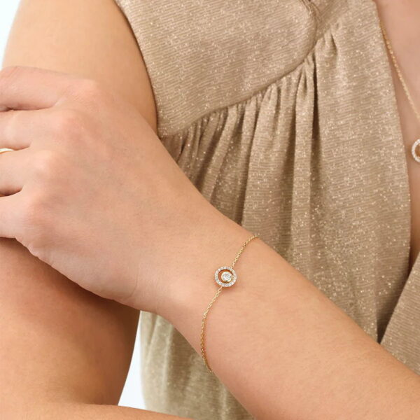 Eine Person trägt das Echtgold Armband Cénacle, ein zartes Goldarmband mit einem runden, diamantbesetzten Anhänger, kombiniert mit einem schimmernden beigen Oberteil. Der Fokus liegt auf dem Handgelenk und dem Schmuck.