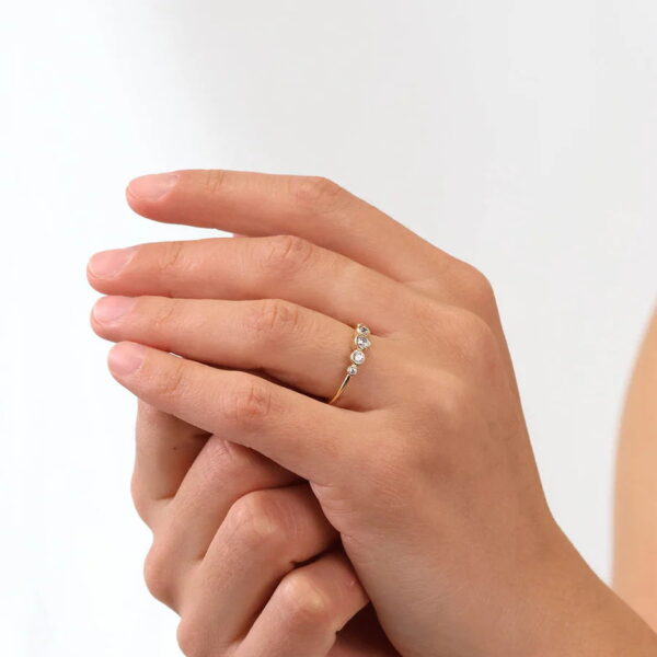 Nahaufnahme einer Hand, die den Echtgold Ring Rangée trägt, ein zartes Goldband mit fünf kleinen runden Diamanten in einer Reihe, gefasst vor einem schlichten weißen Hintergrund.