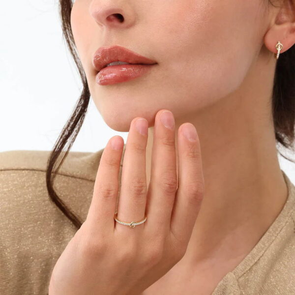 Eine Frau mit glänzenden Lippen berührt ihr Gesicht. Sie trägt den Echtgold Ring Facile - einen Goldring mit einem kleinen Stein - und einen passenden Goldohrring. Sie ist mit einem beigen Oberteil bekleidet, und ihr braunes Haar ist locker frisiert.