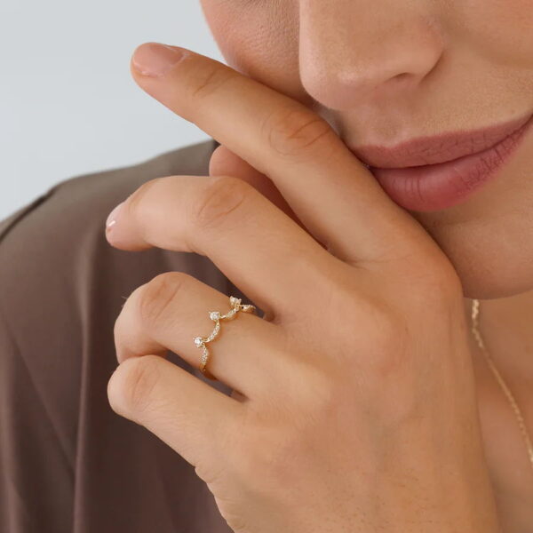 Eine Person mit heller Haut berührt sanft ihr Gesicht. Sie trägt den Echtgold-Ring Princier - einen Goldring mit einem wellenförmigen Muster und kleinen Diamanten. Ihre Lippen zeigen ein leichtes Lächeln und sie trägt braune Kleidung.