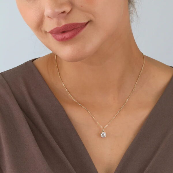 Eine Frau trägt ein braunes Oberteil mit V-Ausschnitt und das Echtgold Collier Èmera, eine zarte goldene Halskette mit einem runden, klaren Edelsteinanhänger. Das Bild ist so beschnitten, dass ihr Gesicht von der Nase abwärts und ihre obere Brust zu sehen sind.