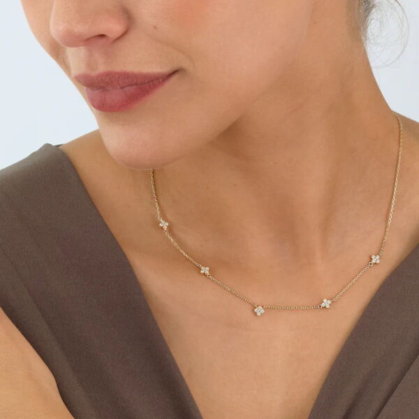 Eine Frau in einem taupefarbenen Kleid mit V-Ausschnitt präsentiert das Echtgold Collier Améline, ein zartes Goldcollier, das mit gleichmäßig verteilten kleinen blütenförmigen Diamanten besetzt ist.